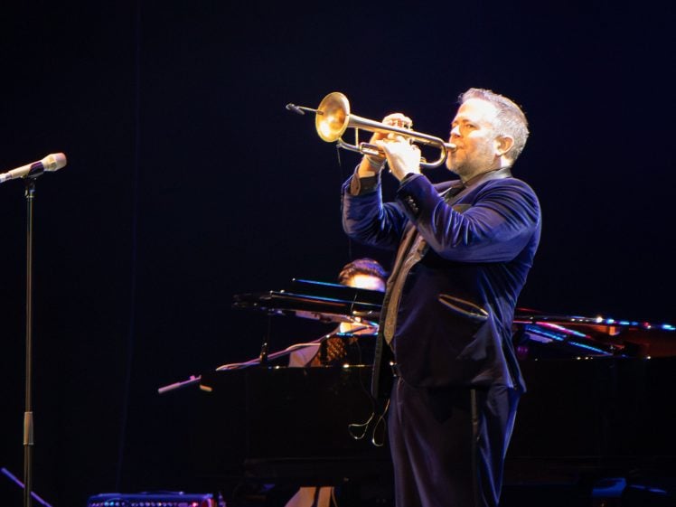 Image of A man in a suit plays the trumpet on stage under bright lights, with a microphone in front of him and a pianist playing on a grand piano in the background.