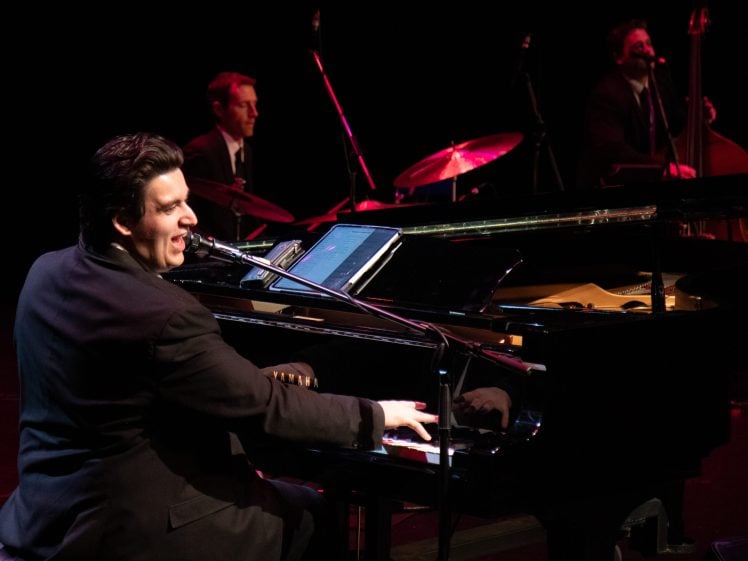 Image of A man in a suit plays a grand piano and sings into a microphone on stage, with a drummer and a bassist performing in the background under stage lighting.