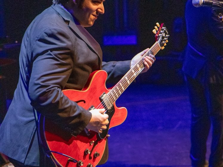 Image of A musician in a black suit plays a red electric guitar on stage under colourful lighting, while another performer is partly visible in the background.