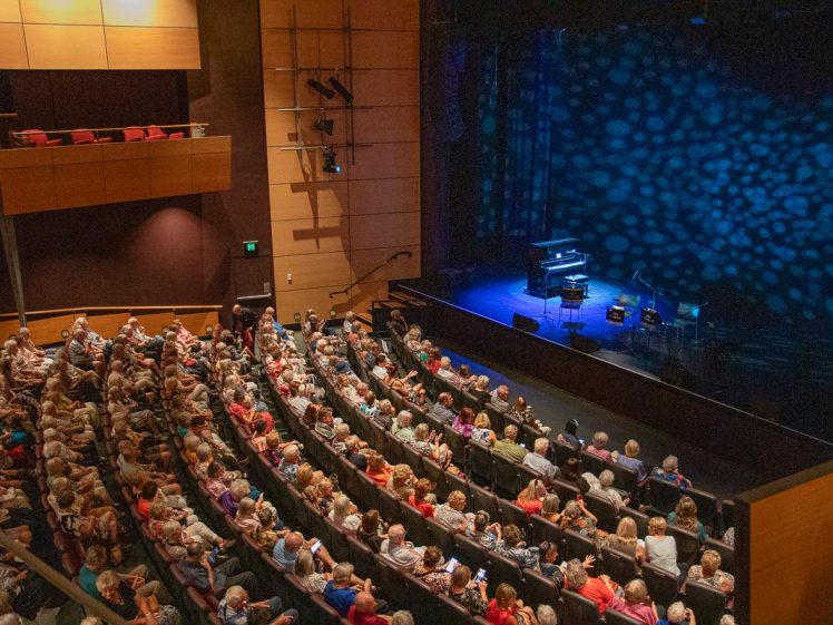 Image of A large audience sits in a modern theatre with curved rows, watching a stage lit in blue with instruments set up, awaiting a performance. Upper balcony seats are visible on the left.