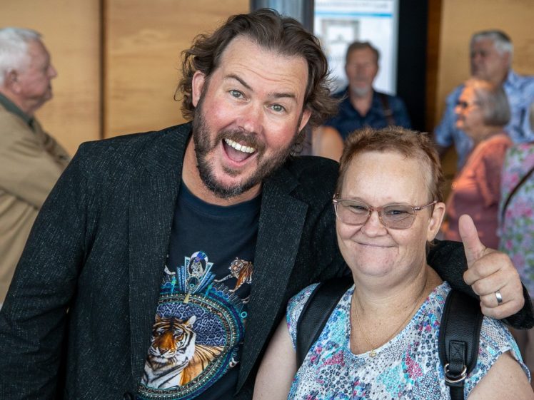 Image of A smiling man with wavy hair and a beard, wearing a black jacket and a tiger shirt, poses with a woman in glasses and a patterned top who gives a thumbs up. Several people are in the background.
