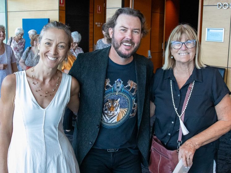 Image of Three smiling adults pose together indoors at an event. The man in the middle wears a black blazer and a graphic T-shirt, while the two women on either side wear light-coloured tops. People are gathered in the background.