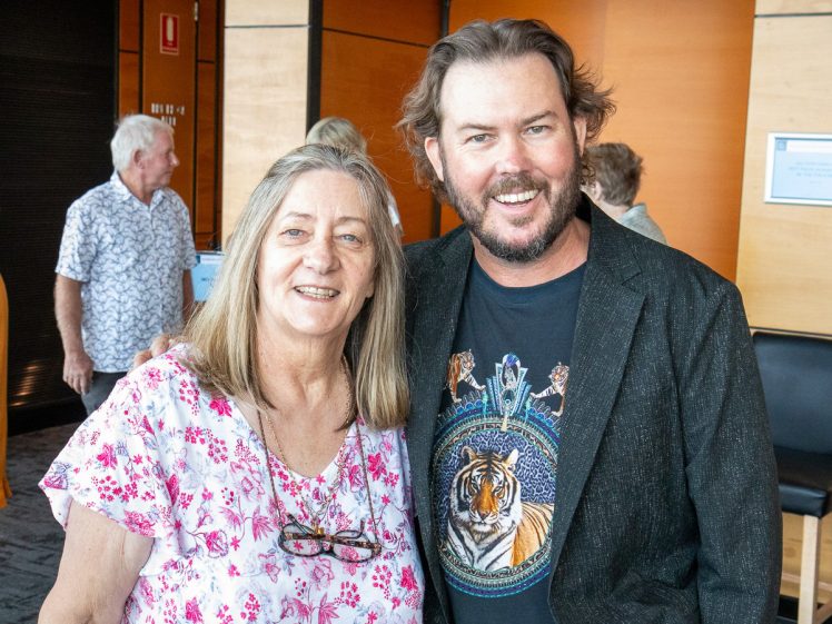 Image of A woman in a floral shirt and a man in a black jacket and tiger T-shirt smile together indoors, with other people and wood-panelled walls in the background.