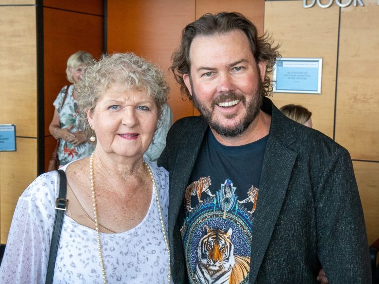 Image of An older woman with curly grey hair and a younger man with wavy brown hair and a beard stand smiling together indoors. The man wears a black blazer and tiger-themed shirt; the woman wears a light-patterned top and pearl necklace.