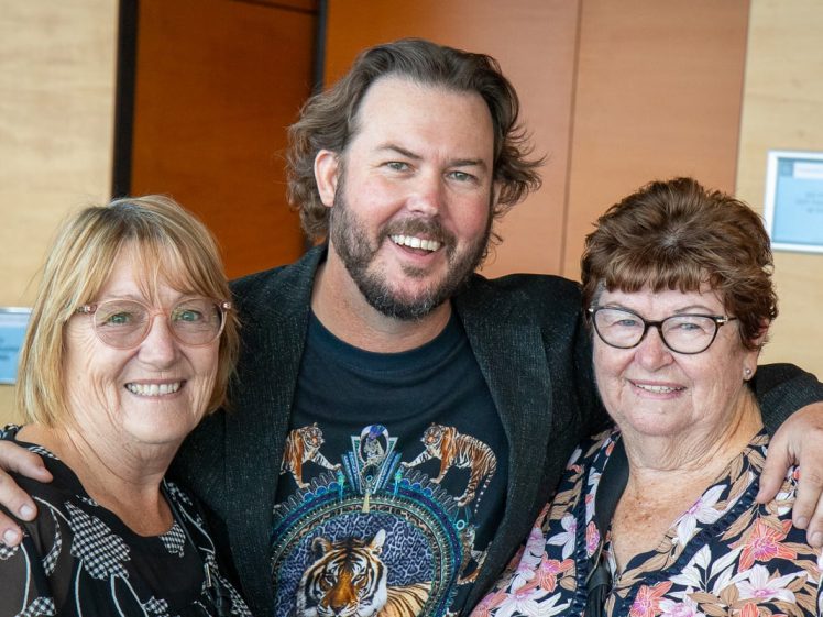 Image of A smiling man with a beard stands between two older women, all with their arms around each other, posing happily indoors. The women wear glasses and floral tops; the man wears a dark jacket and a T-shirt with tiger designs.