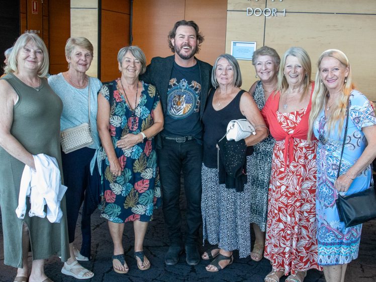 Image of A group of eight smiling adults, one man and seven women, pose together indoors near a wooden wall and a door labelled DOOR 1. The group is casually dressed and standing close to each other.