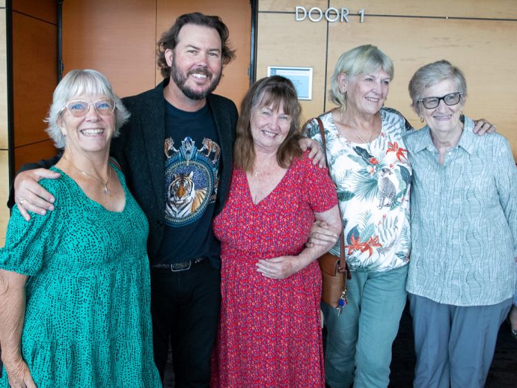 Image of Five people stand smiling together indoors. Four women, wearing glasses and colourful outfits, stand with a man in a black blazer and graphic shirt. They pose closely in front of a wood-panelled wall marked Door 1.