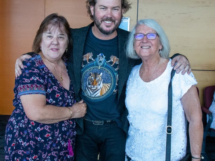 Image of Three people stand smiling together indoors. The man in the middle has his arms round two older women, one wearing a patterned dress and the other in a white blouse and glasses. They appear happy and posing for a photo.