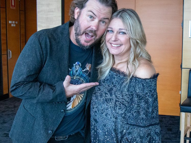 Image of A man with a beard and a casual blazer enthusiastically points to a smiling woman in a patterned off-the-shoulder dress as they pose together indoors against a wooden wall.