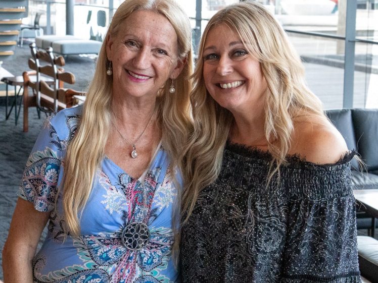 Image of Two smiling women with long blonde hair stand close together indoors. One wears a blue patterned dress and necklace; the other wears a dark off-the-shoulder top. Modern lounge furniture and large windows are in the background.