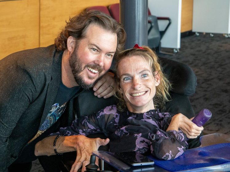 Image of A man with a beard leans in, smiling, next to a woman with a disability in a wheelchair. The woman is also smiling and holding a purple object; both appear happy in a casual indoor setting.