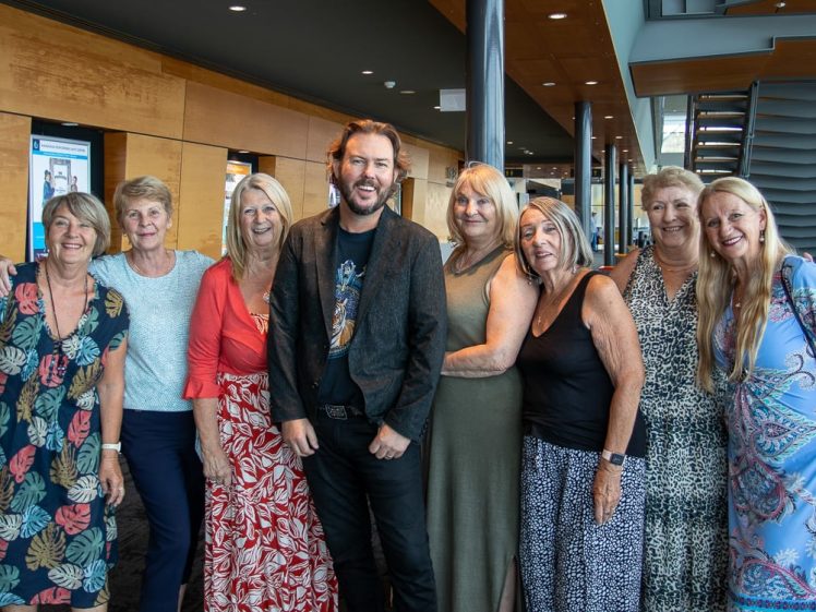 Image of A group of eight people, seven women and one man, stand indoors smiling together. They are dressed in colourful and casual clothing, with wood panelled walls and a staircase in the background.