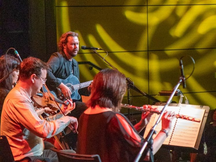 Image of A guitarist sings into a microphone on stage, accompanied by musicians playing violin and flute, with yellow patterned stage lights projected on the wall behind them.