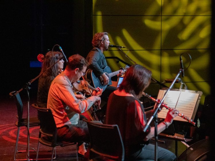 Image of Four musicians perform on stage: one sings and plays guitar, while the others play string and wind instruments. They sit in a row with music stands, illuminated by stage lights and projected patterns on the wall behind them.
