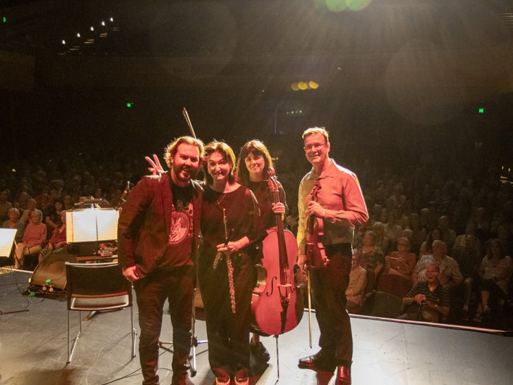 Image of Four musicians stand on a stage smiling at the camera, holding their instruments, with bright stage lights shining above and an audience seated in the dark background.