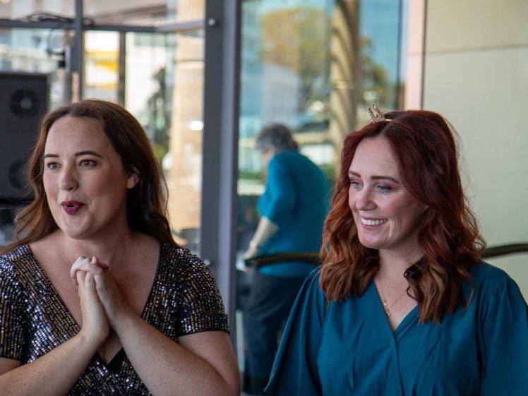 Image of Two women stand indoors, one in a sparkly dress speaking with hands clasped, and the other in a teal dress and a small crown, smiling. A reflection of a person is visible in the glass behind them.