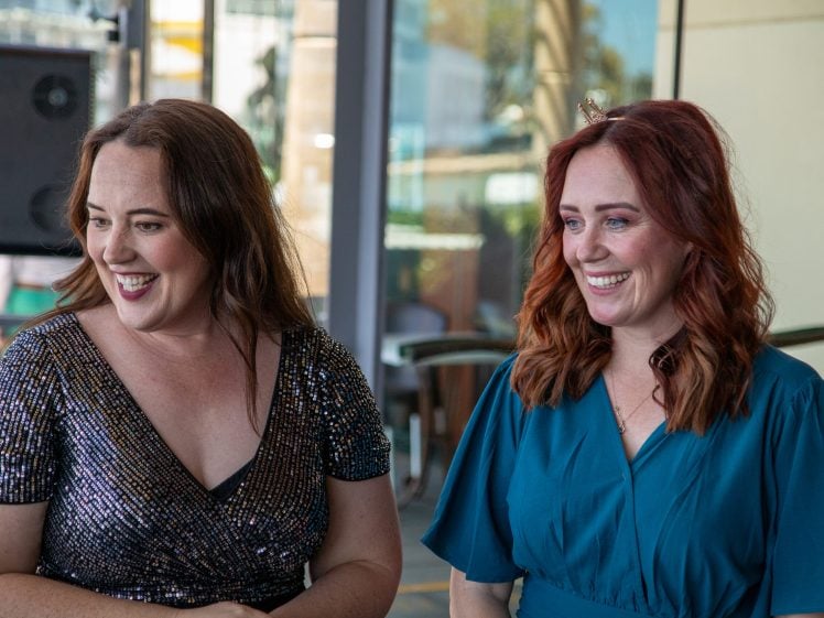 Image of Two women with wavy brown and red hair are smiling and standing side by side indoors. One wears a sparkly black dress and the other a blue dress with a small hair accessory. Glass windows and chairs are in the background.