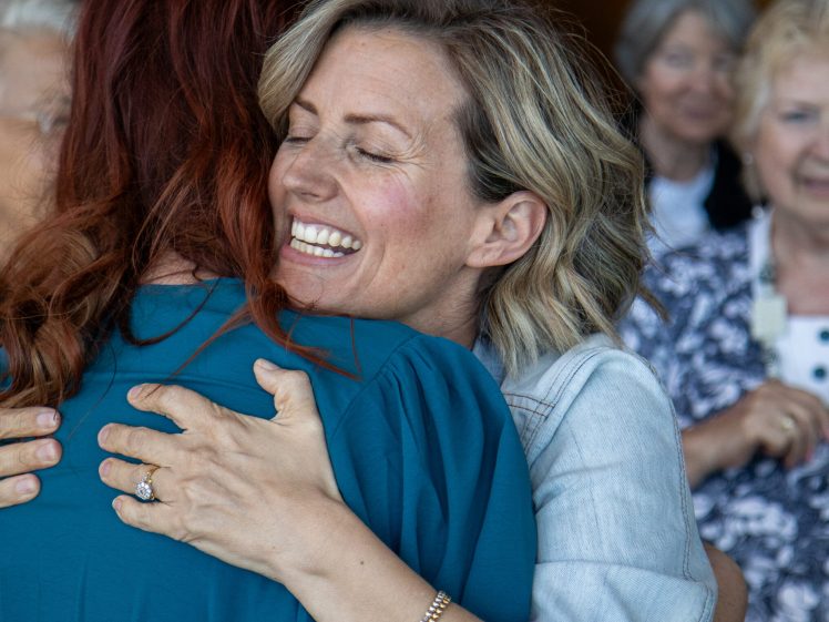 Image of A smiling woman with blonde hair embraces another woman with long red hair, whilst people in the background look on and smile. The atmosphere appears warm and joyful.