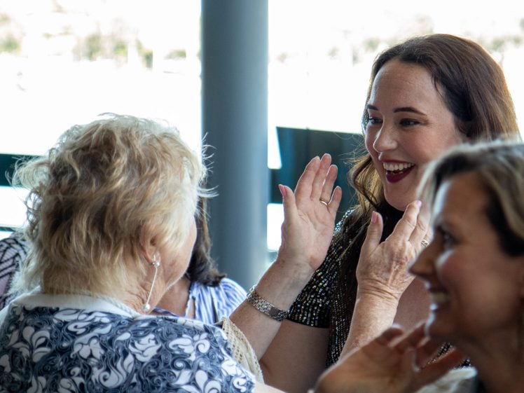 Image of Two women smiling and talking animatedly at an indoor gathering, with one woman raising her hands in excitement. Other people are visible in the foreground and background.