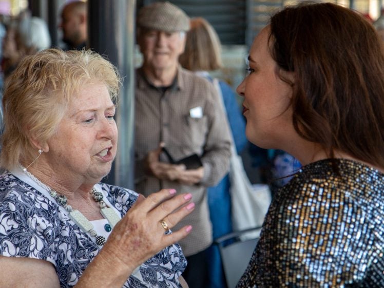 Image of An older woman gestures while talking to a younger woman in a sequinned top at a social gathering. People are visible in the background, including a man wearing a flat cap and holding a notebook.