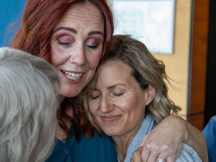 Image of Two women are hugging closely, both with content, peaceful expressions. One woman has red hair and closed eyes; the other, with blonde hair, is smiling softly with her eyes closed. A third person is partly visible in the foreground.