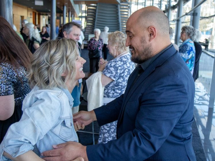 Image of A man in a dark suit and a woman in a light denim jacket greet each other warmly, smiling and holding each other's arms, in a bright indoor space with several other people in the background.