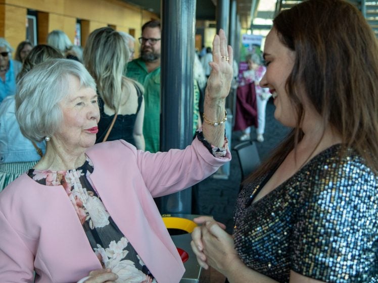 Image of An elderly woman in a pink jacket playfully reaches towards a smiling younger woman in a sparkly dress at a lively indoor gathering, with other people socialising in the background.