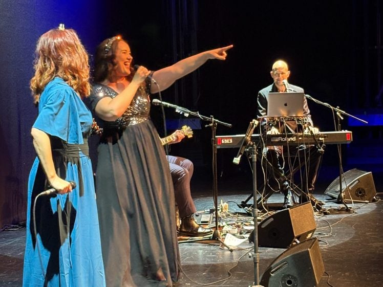 Image of Two women in elegant dresses perform on stage, one singing and pointing into the audience. A man seated at a keyboard and microphone plays music. Stage lights and musical equipment are visible in the background.