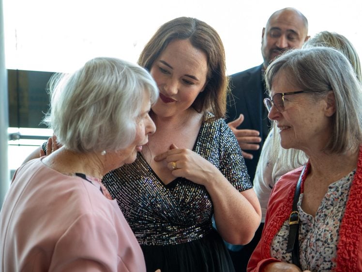 Image of Three women stand together, smiling and talking at an indoor event. One woman in a sparkly dress listens attentively as the other two, both with grey hair, chat closely. A man in a suit is visible in the background.