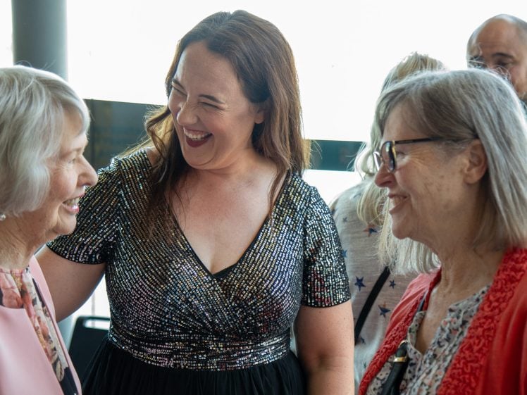 Image of Three women smile and chat together at an indoor event, dressed in semi-formal attire. The woman in the centre wears a sparkly black dress, while the others wear glasses and light-coloured tops. Other people are blurred in the background.
