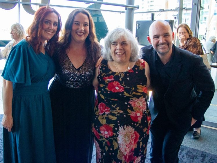 Image of Four adults stand close together, smiling for the camera at an indoor event. Two women wear dresses—one teal, one floral—whilst the other woman wears a sparkly black dress. The man wears a black suit. Other people are visible in the background.