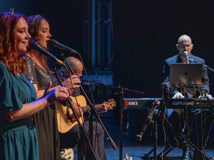Image of Three musicians perform on stage: two women sing into microphones, one in a blue dress and the other in a sparkling top, while a man plays keyboard and another plays guitar in the background. Stage lighting is dim.