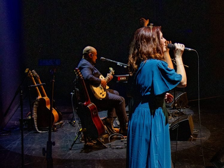 Image of A woman in a blue dress sings into a microphone on stage whilst a man sits and plays guitar. Musical equipment and two acoustic guitars are visible in the dimly lit background.