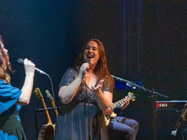 Image of Two women sing passionately on stage under dramatic lighting, one facing the audience and one in profile. A man plays a keyboard and sings in the background. Musical instruments and equipment are visible.