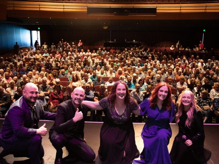 Image of Five performers pose and smile at the front of a theatre stage, facing the camera, with a large audience seated behind them in a well-lit auditorium.