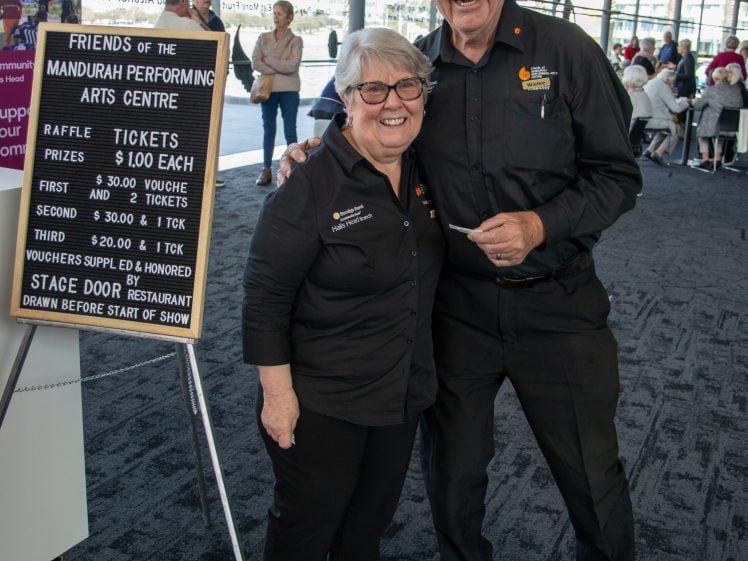 Image of Two older adults in black uniforms smile and pose together beside a raffle sign for the Mandurah Performing Arts Centre at an indoor event with people seated in the background.