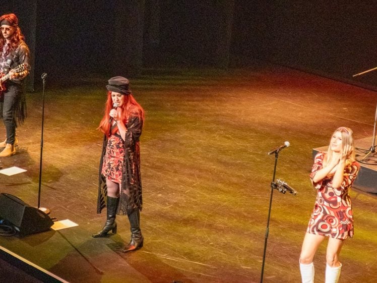 Image of Three musicians perform on stage under warm lighting; one plays guitar, one sings at centre wearing a hat, and one stands near a microphone in a patterned dress and white boots, hands pressed together.