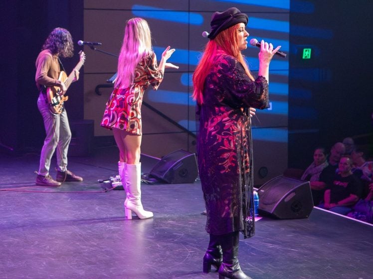 Image of Three musicians perform on stage: a guitarist, a singer with long blonde hair in a retro dress and white boots, and a red-haired singer in a black outfit and hat, with an audience watching from the right.