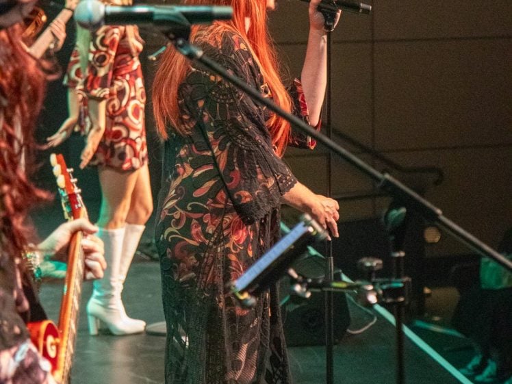 Image of A woman with long red hair, wearing a black hat and patterned dress, sings into a microphone on stage. Other musicians and a woman in a retro outfit with white boots are also performing. Stage lights illuminate the scene.