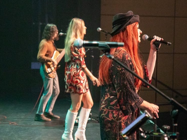 Image of Three musicians perform on stage. A woman with long red hair sings into a microphone, another woman with blonde hair sings beside her, and a guitarist plays in the background. Stage lights illuminate the scene.