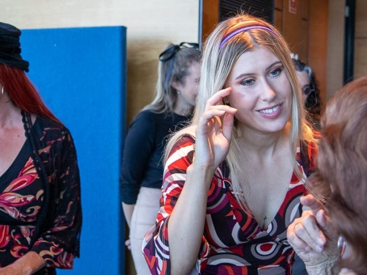 Image of Two women in patterned dresses smile and talk at an indoor event. One woman with blonde hair gestures with her hand near her face, whilst another red-haired woman stands nearby, also smiling. Other people are in the background.
