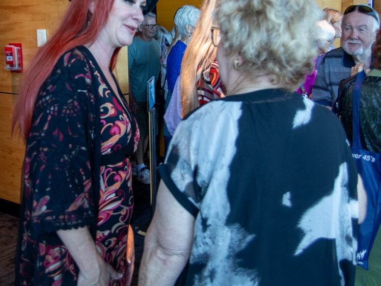 Image of A smiling woman with long red hair and a black hat talks to an older woman with blonde hair and glasses in a crowded indoor space with wood-panelled walls. Other people mingle in the background.