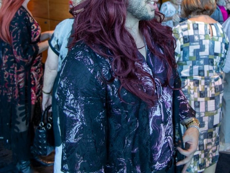 Image of A man in a shiny purple, patterned shirt, black lace jacket, sunglasses, and a long burgundy wig stands in a crowded indoor space, talking to others. Several people are gathered in the background, some with hats.