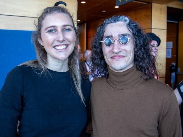 Image of Two people pose and smile for a photo indoors. The person on the left has long light brown hair and wears a black top, whilst the person on the right has curly dark hair, round glasses, and a brown polo neck.