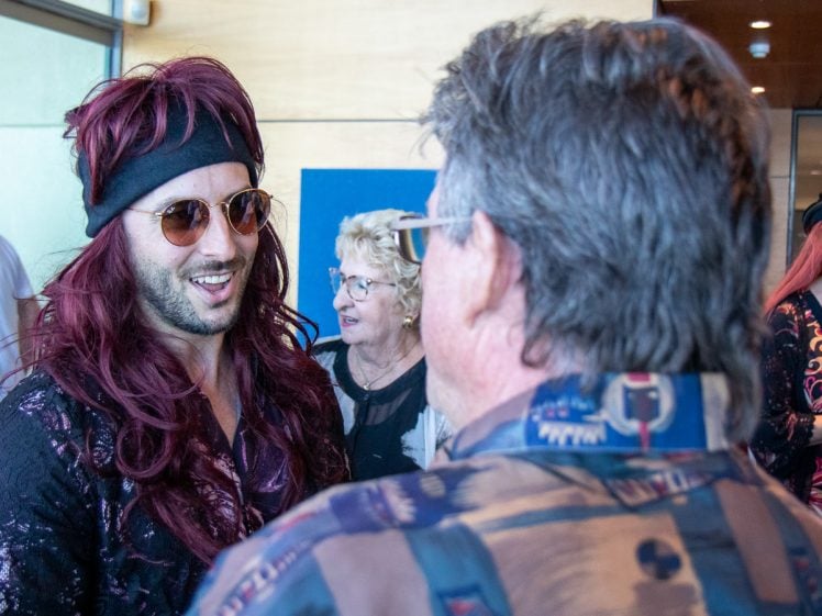 Image of A man with long red hair, sunglasses, and a black headband smiles whilst talking to another man in a patterned shirt. A woman with blonde hair stands in the background, looking on.