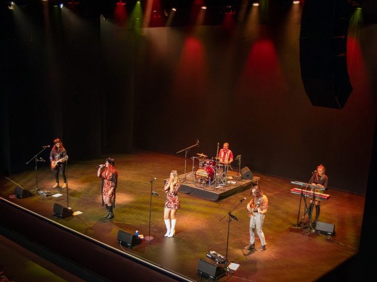 Image of A band of six musicians performs on stage under colourful lights. One plays drums, one is at a keyboard, and the others play guitars or sing into microphones. The stage is dark with red, yellow, and green spotlights overhead.