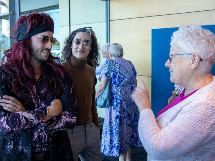 Image of A woman with short white hair and glasses talks to two men; one wears a long red wig, headband, and sunglasses, while the other has curly hair and round glasses. People in patterned blue dresses stand in the background.