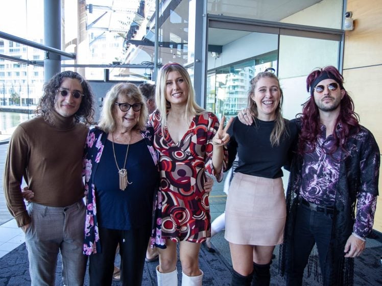 Image of Five people stand together indoors, smiling at the camera. Three are dressed in vintage or retro-style clothing, including patterned dresses, a long cardigan, sunglasses, and a hairband. The setting is modern with glass walls.