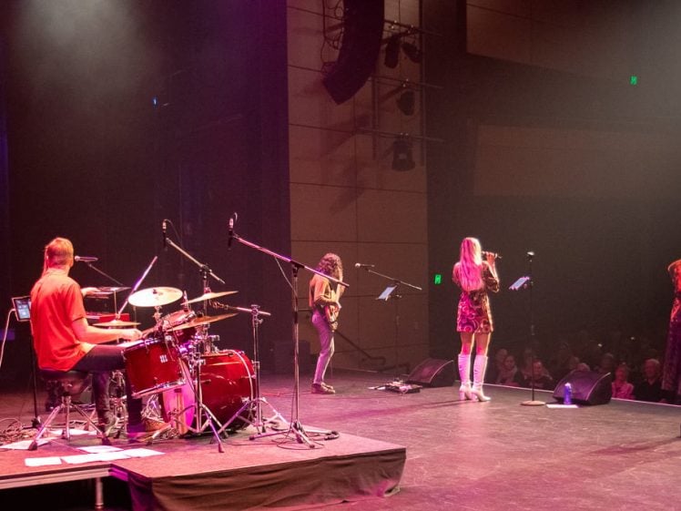 Image of A band performs onstage with a keyboard player, drummer, guitarist, and two vocalists under colourful lights, facing an audience in a dimly lit theatre.