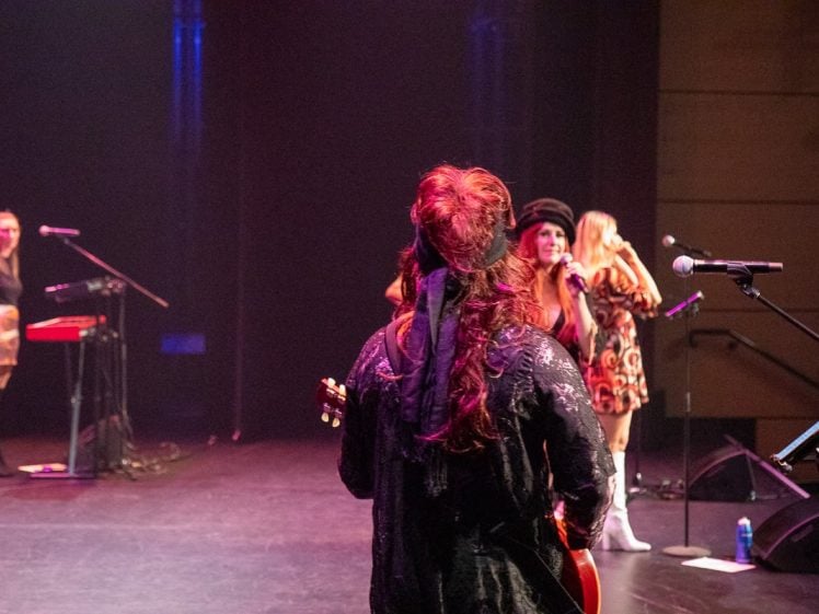 Image of A musician with long hair stands on stage facing three performers near microphones; one plays keyboard and two sing. Stage lighting is dim, with blue and red hues, and a water bottle is on the floor.